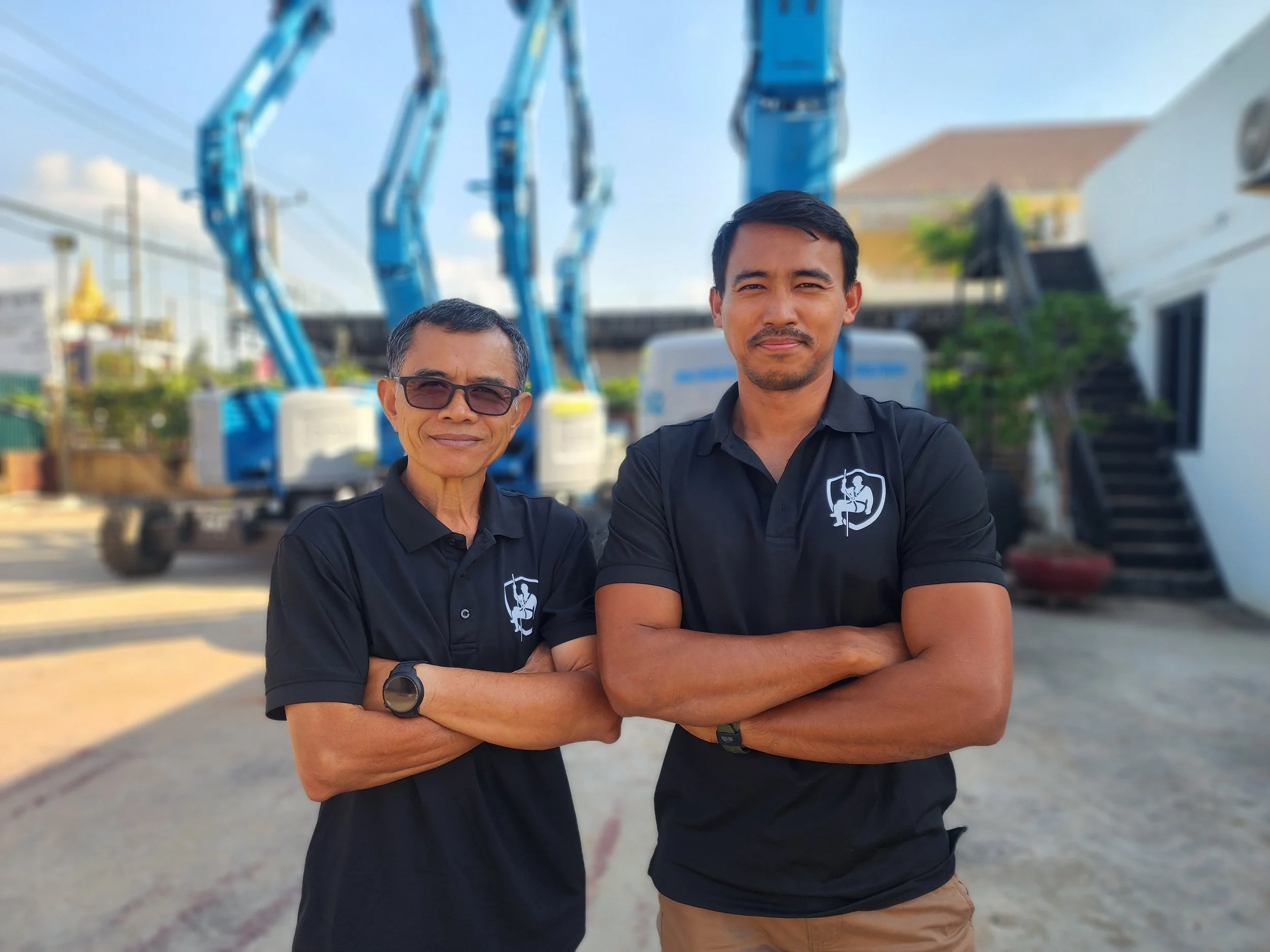 Two men standing outdoors in front of blue construction lifts, both wearing black shirts with a logo on the left chest, arms crossed, smiling at the camera.