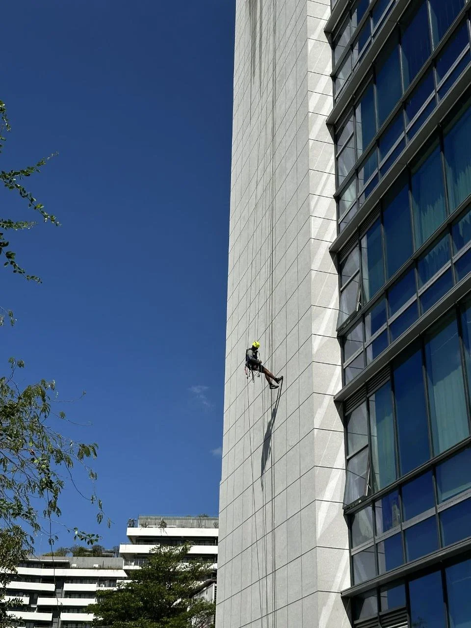 A worker wearing a helmet and safety harness is cleaning the side of a tall modern office building using rope access techniques.