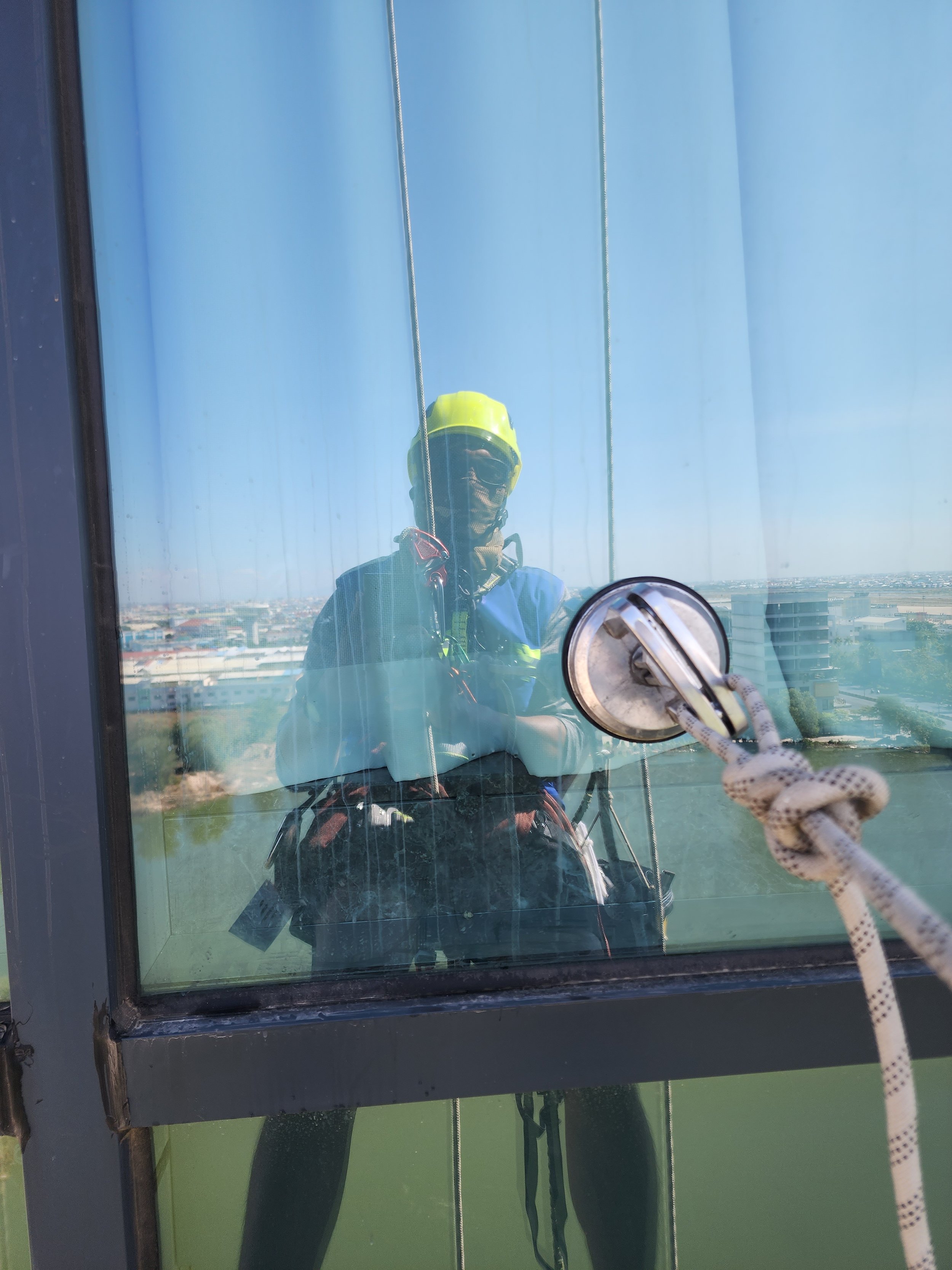 Person cleaning a tall glass window from the outside, wearing safety gear including a helmet, goggles, and harness, using a cleaning tool attached to a rope.
