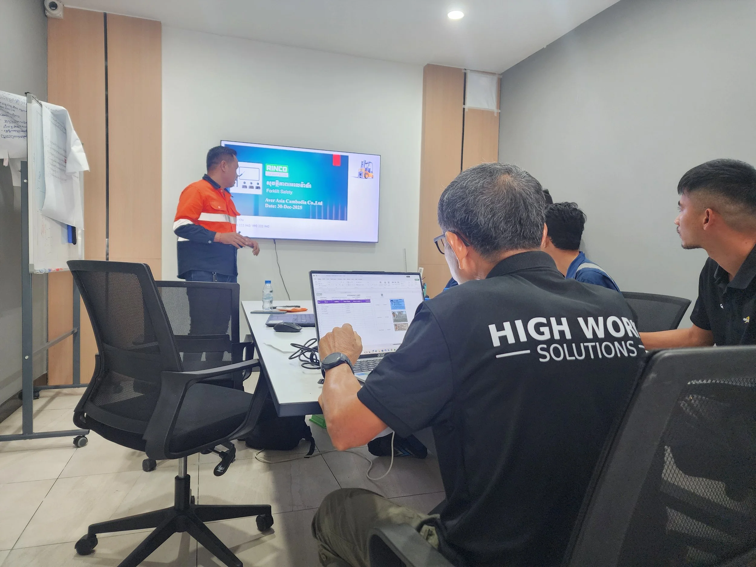 A man giving a presentation on forklift safety to a group of five men in a conference room. One man is wearing a black shirt with 'High Works Solutions' written on the back. The others are listening attentively. There is a large screen displaying a slide with blue, green, and red colors, and a water bottle on the table.