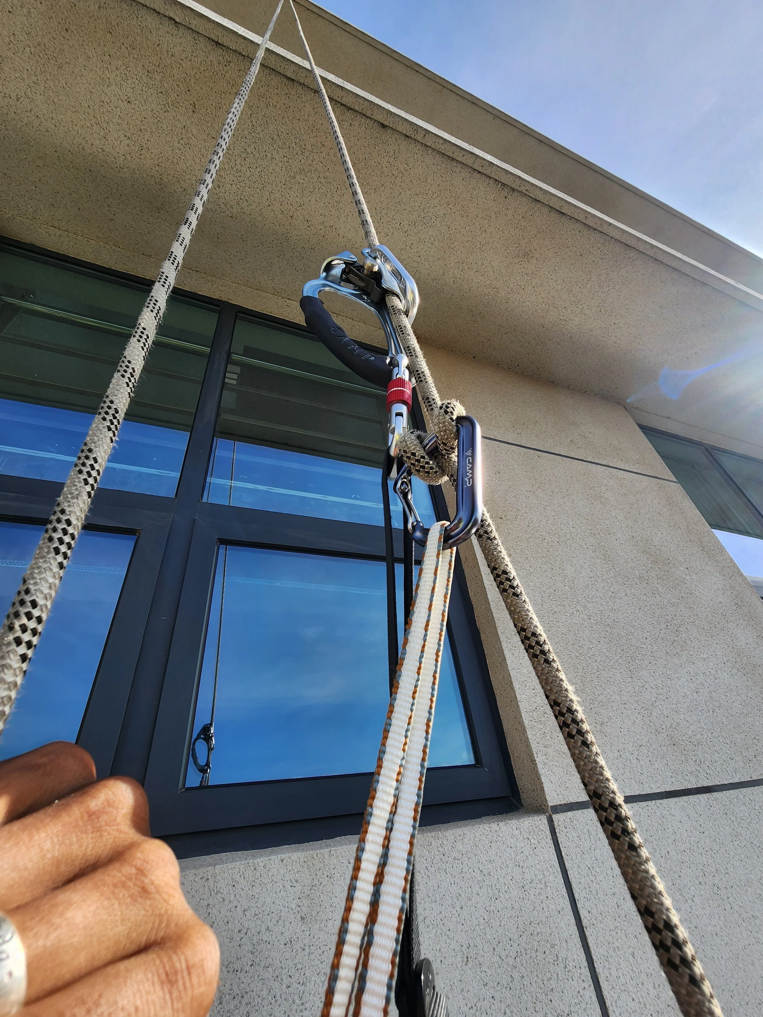 Worker using climbing gear with ropes and carabiners on a building exterior to clean or maintain a window.