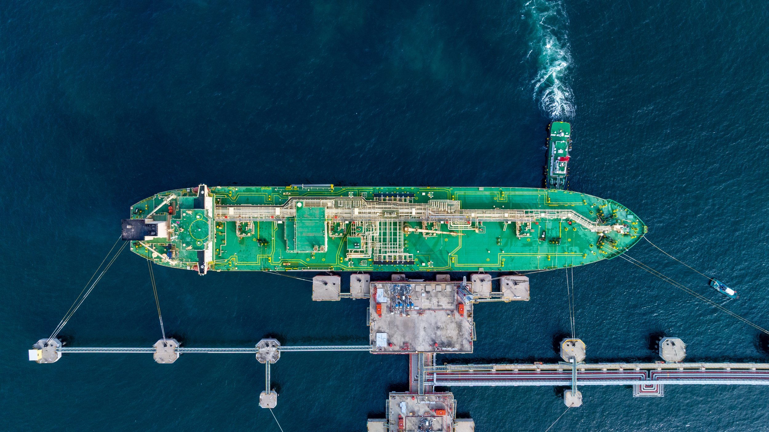 An aerial view of a large oil tanker ship docked at an offshore platform or oil rig at sea. The ship's deck is covered with green-colored piping and equipment.