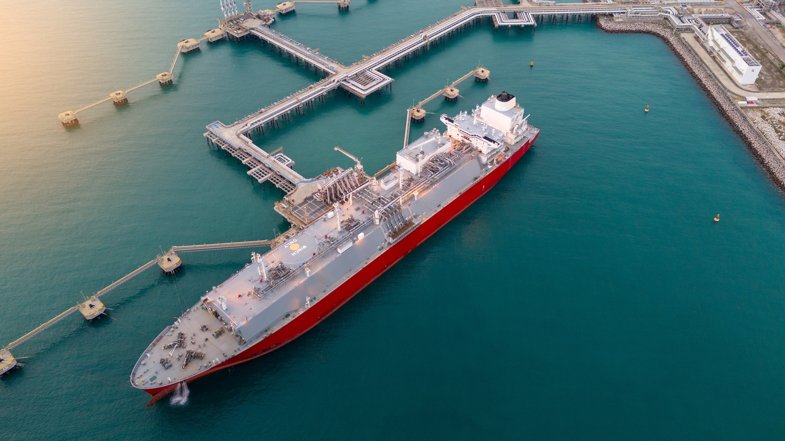 An aerial view of a large cargo ship docked at a pier, with water and a breakwater wall nearby.