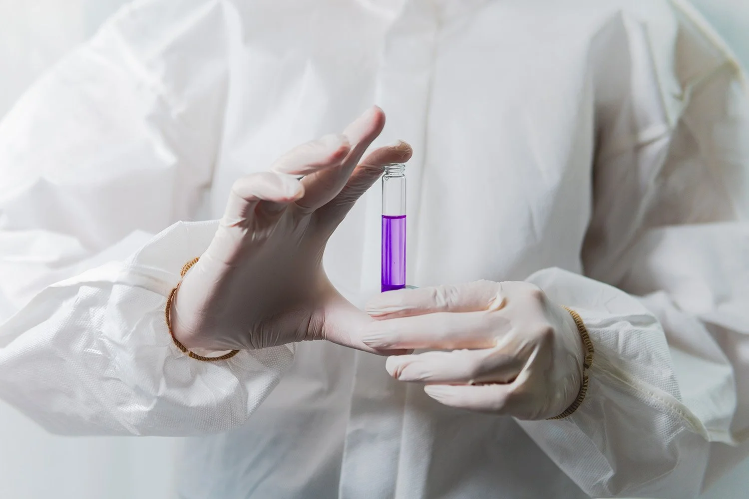 Person wearing white protective lab coat and gloves holding a small test tube with purple liquid.