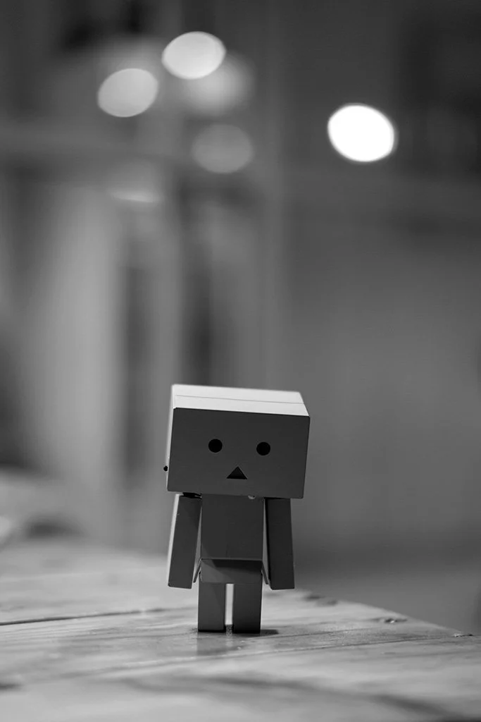 A small cardboard robot figure with a square head and simple facial features standing on a wooden surface, with a blurred background featuring lights.