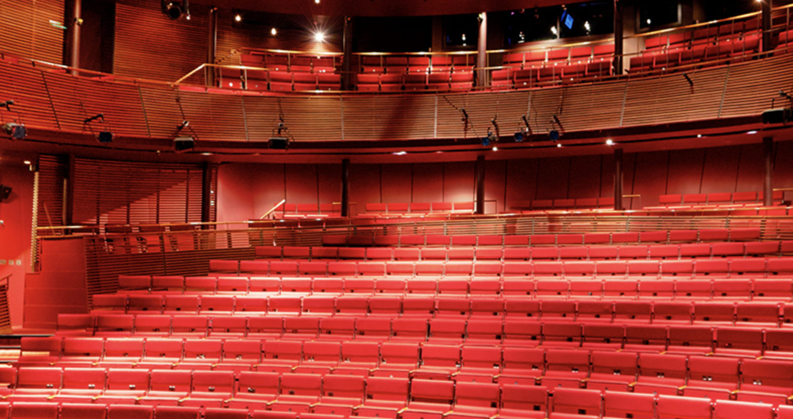 The interior of a theater or auditorium with red seats and wooden accents, shown from the stage area looking towards the audience.