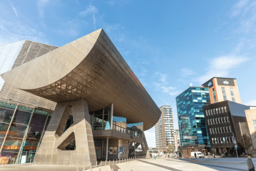 Modern cityscape featuring a large, uniquely designed building with a curved, overhanging roof supported by angled pillars, surrounded by tall glass office buildings.