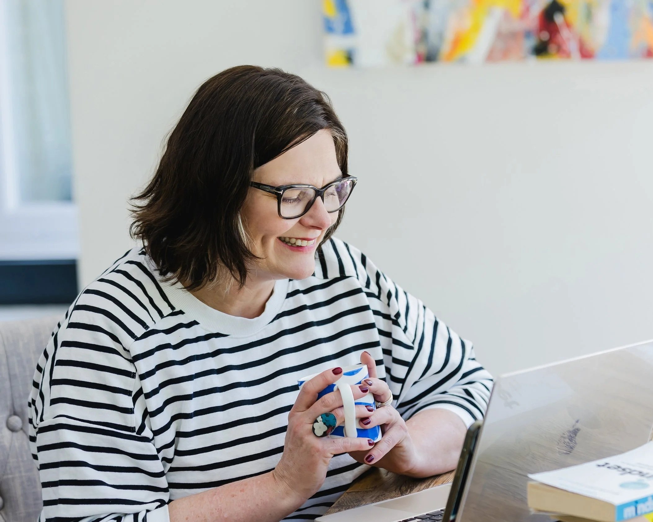 A woman with shoulder-length brown hair, glasses, and a striped black and white shirt is smiling while holding a colorful gift box with a white ribbon, sitting at a table in a bright room.