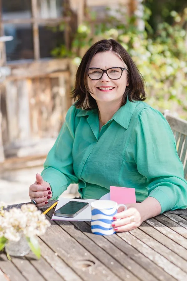 A woman, arts and culture consultant, Jane Macpherson, with glasses and shoulder-length dark hair, wearing a green shirt, smiling and sitting at an outdoor wooden table with a smartphone, a mug, a notepad with a pink sticky note, and a pencil.