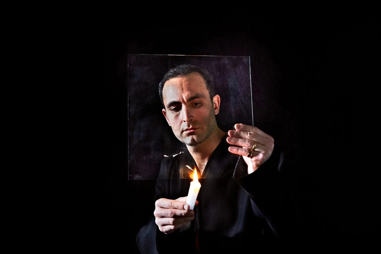 A man looks at a candle flame through a transparent square set against a dark background.