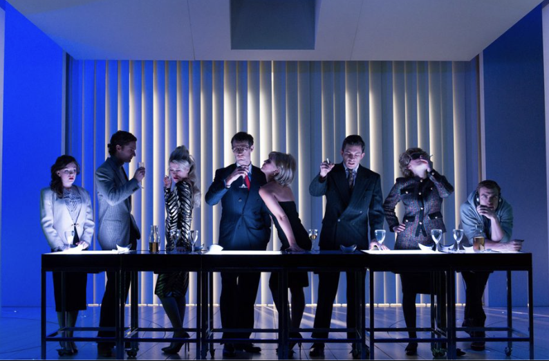 A group of seven people in formal attire gathered around a dining table in a modern, glowing room with vertical light panels, some engaging in conversation, others looking away or deep in thought.