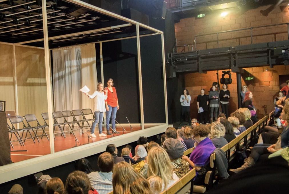 Two women standing on stage for a rehearsal or performance in front of an audience, with chairs and a curtain behind them, and people standing and sitting in the auditorium.