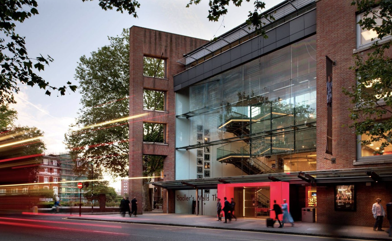 Modern multi-story building with large glass windows, red brick exterior, and a red entrance, with people walking on the sidewalk at dusk.