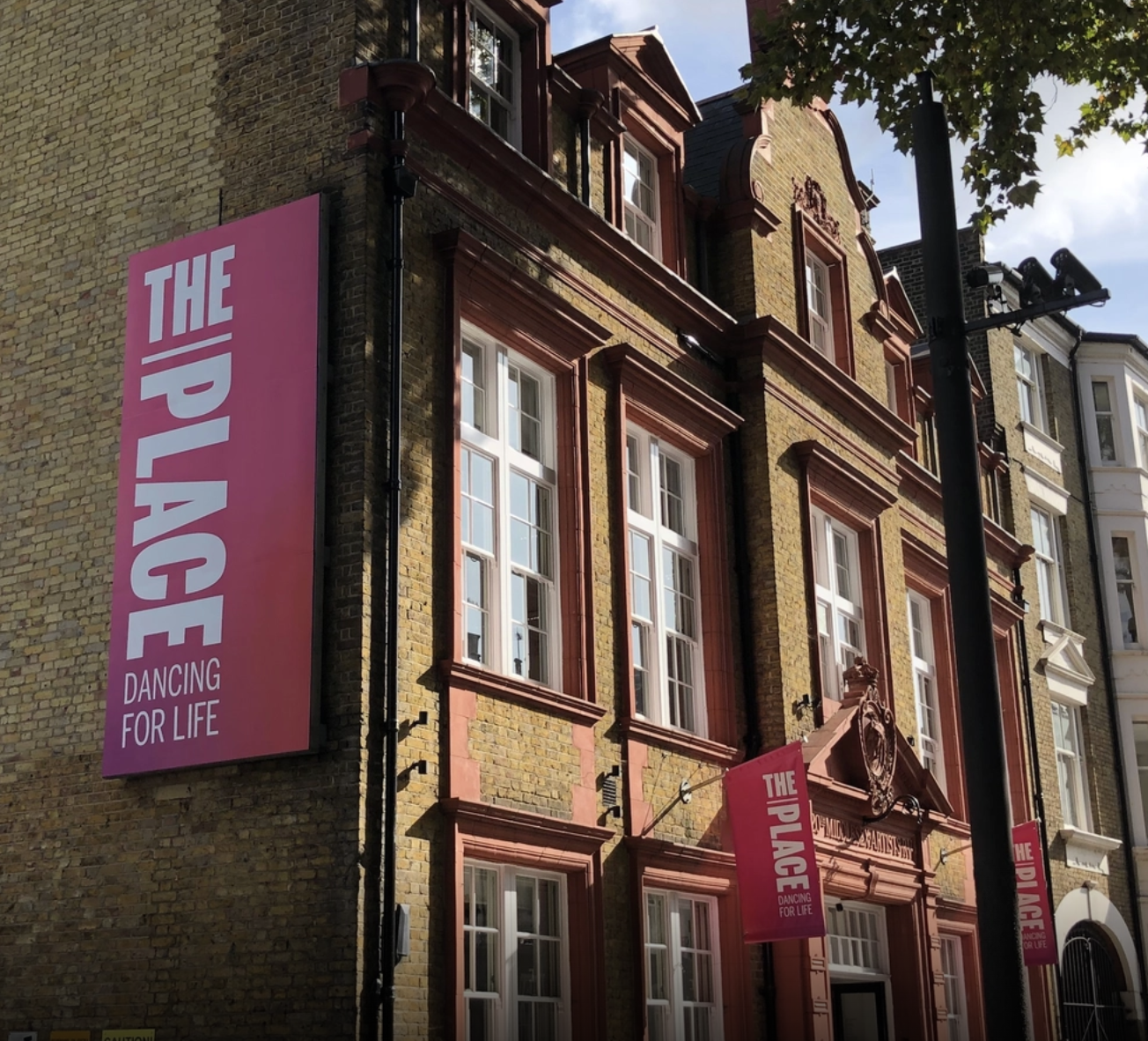 Brick building with multiple white-framed windows and pink banners that read 'The Place Dancing for Life' hanging on the exterior wall.