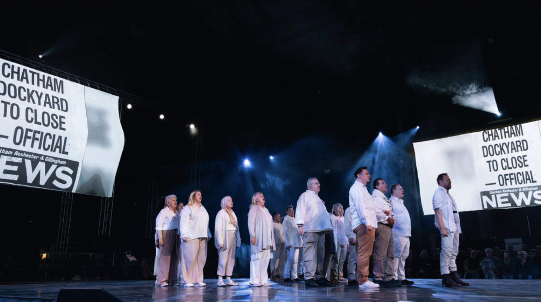 A group of people standing on stage, appearing to be part of a formal event or performance, with large screens displaying the text 'Chatham Dockyard to Close Official News' behind them. The stage is lit with blue and white lights.