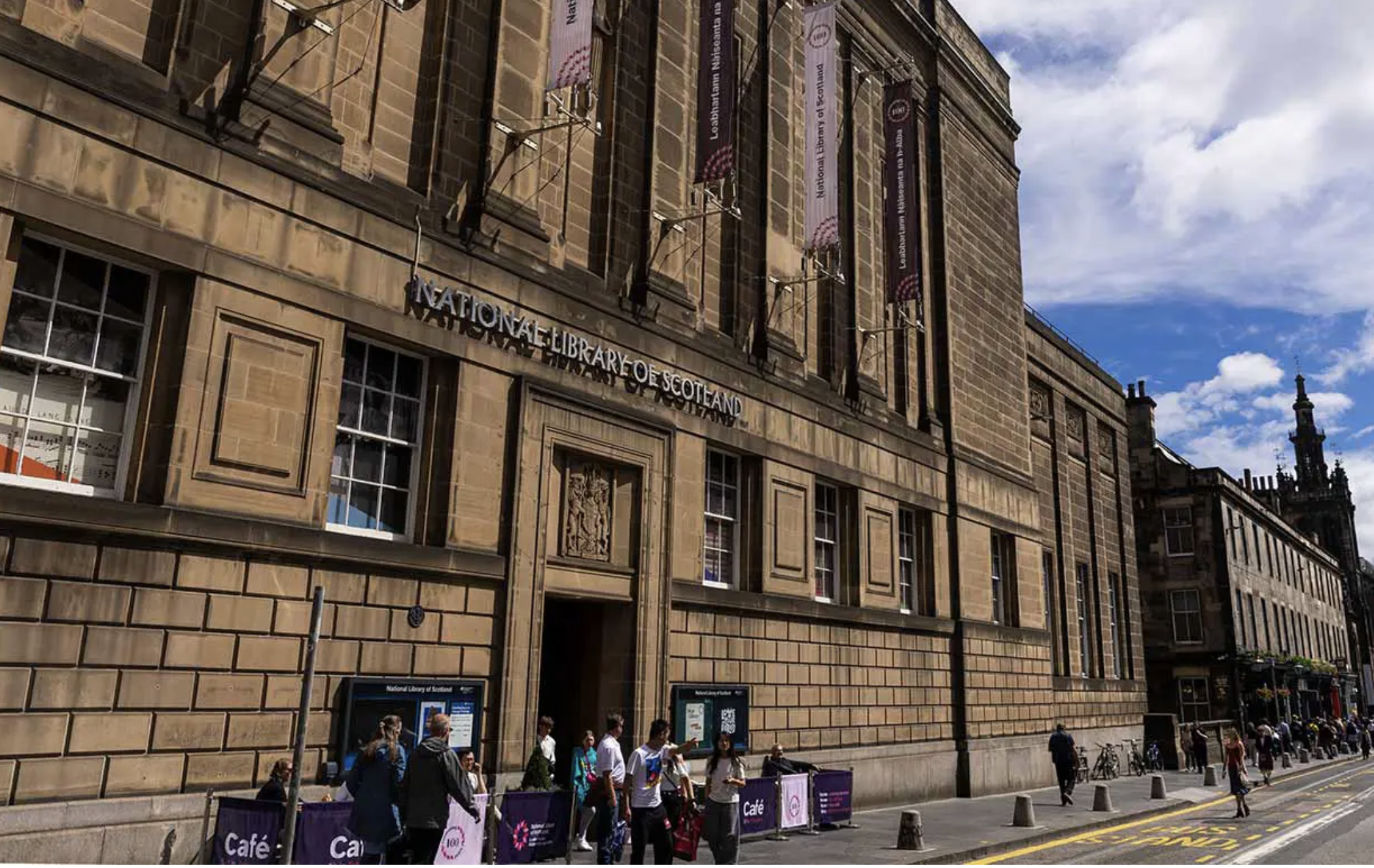 The exterior of the National Library of Scotland building made of stone, with banners hanging from it, and a crowd of people walking on the street underneath.