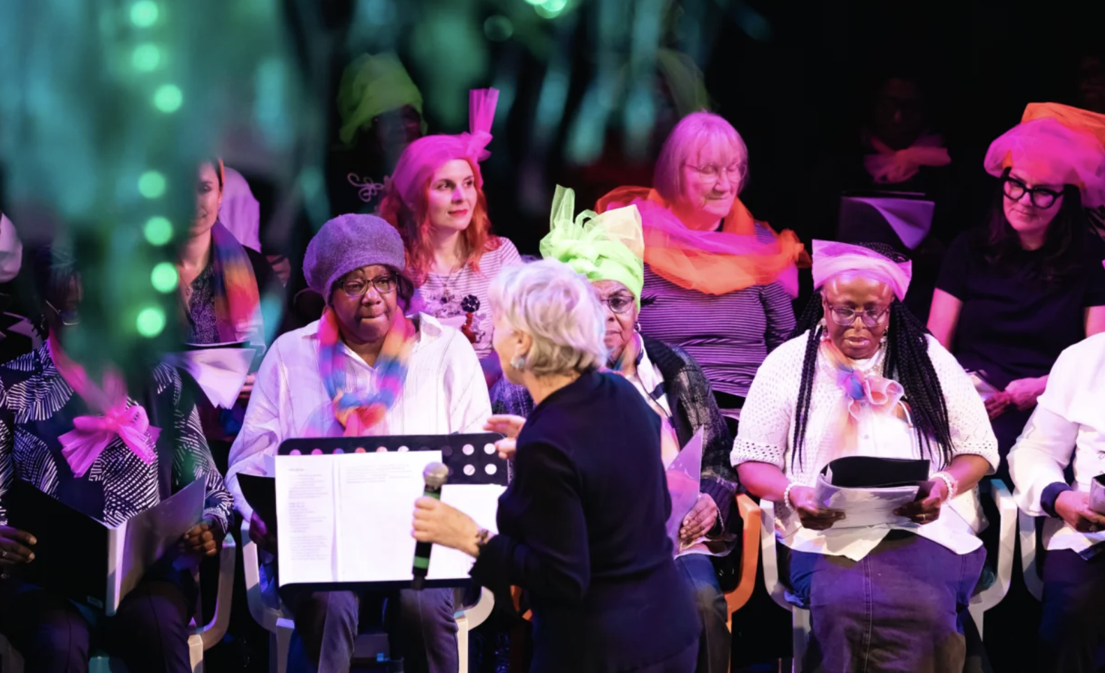 A choir of women, diverse in age and ethnicity, sitting on stage during a performance, wearing colorful headscarves and accessories, while a woman holding a microphone and sheet music addresses them.
