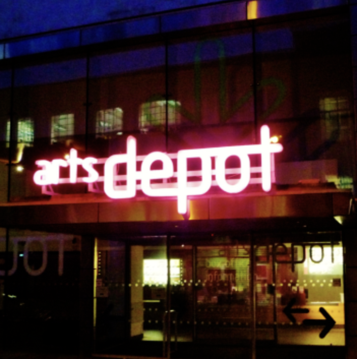 Neon sign that says "arts depot" in pink and white lights, with reflections and windows in the background.