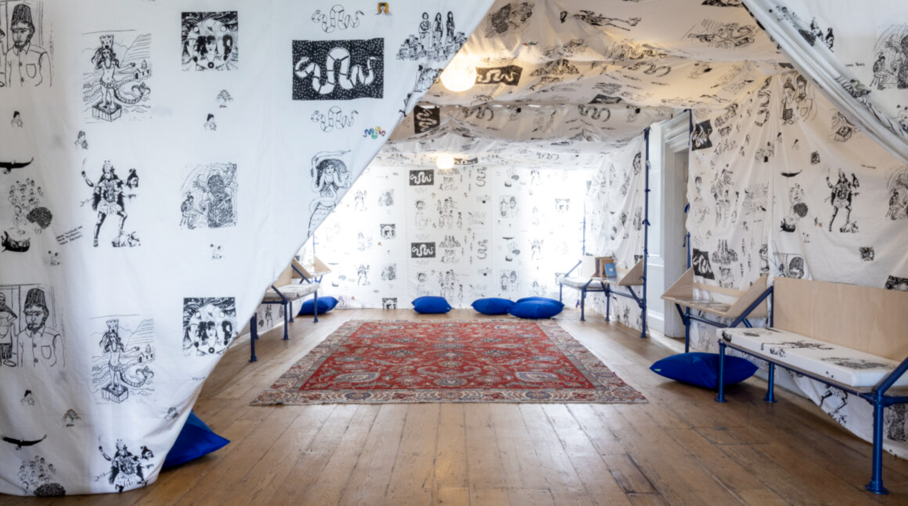 A room with walls and ceiling covered in white paper with black line drawings of various figures, animals, and abstract designs. The floor has a red patterned rug with blue cushions and pillows scattered along the walls. Wooden benches and small tabl