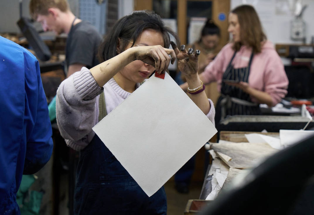 A woman is holding a square piece of white material in a workshop or studio, inspecting it closely amid other people engaged in various activities.