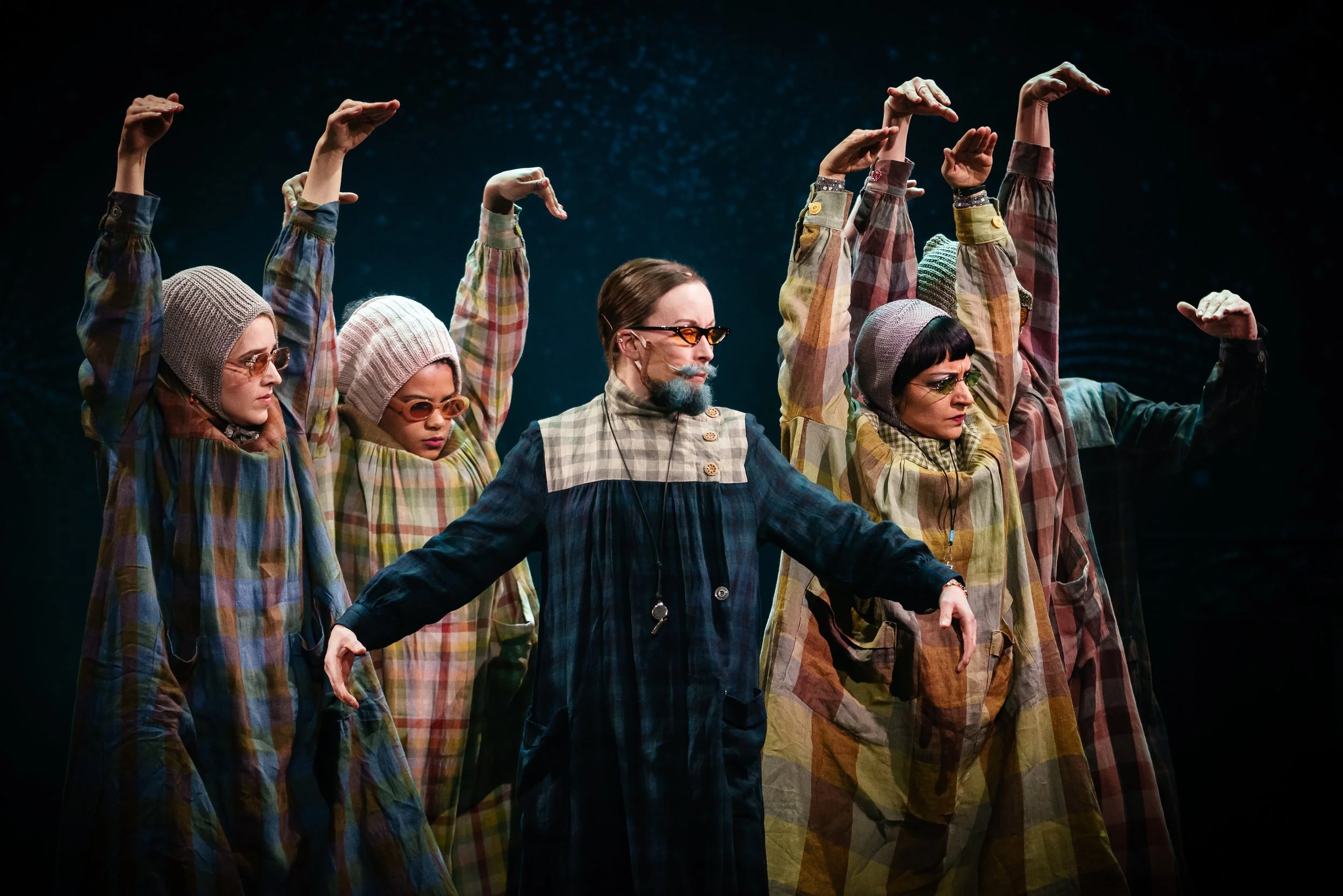 A group of five people in colorful, vintage-style clothing and glasses are performing in a dimly lit space, with their arms raised in a coordinated pose.