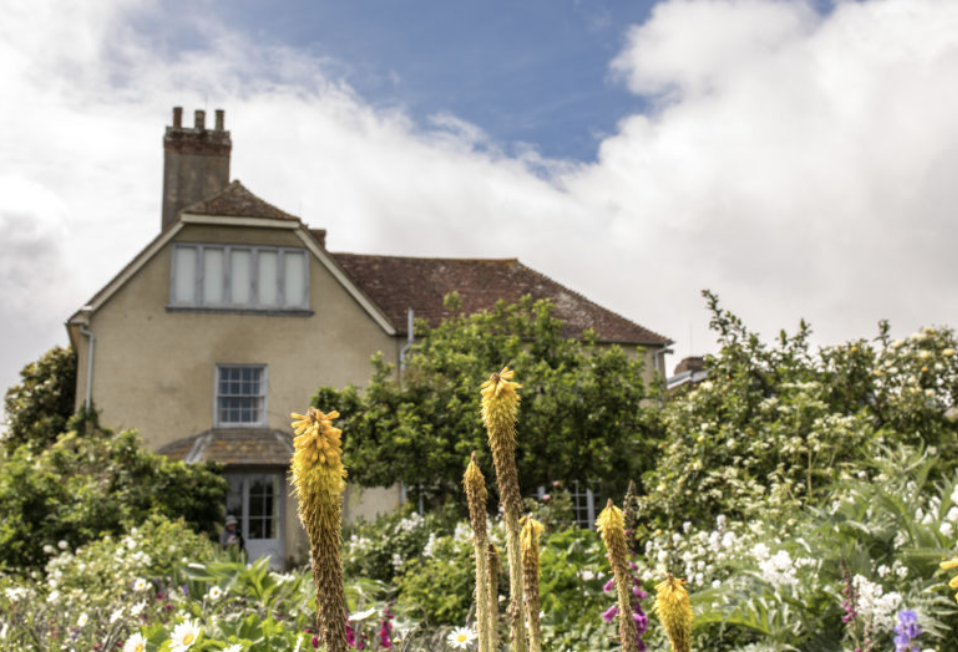 A house with a sloped roof and chimney behind a lush garden filled with colorful flowers and green bushes, under a partly cloudy sky.