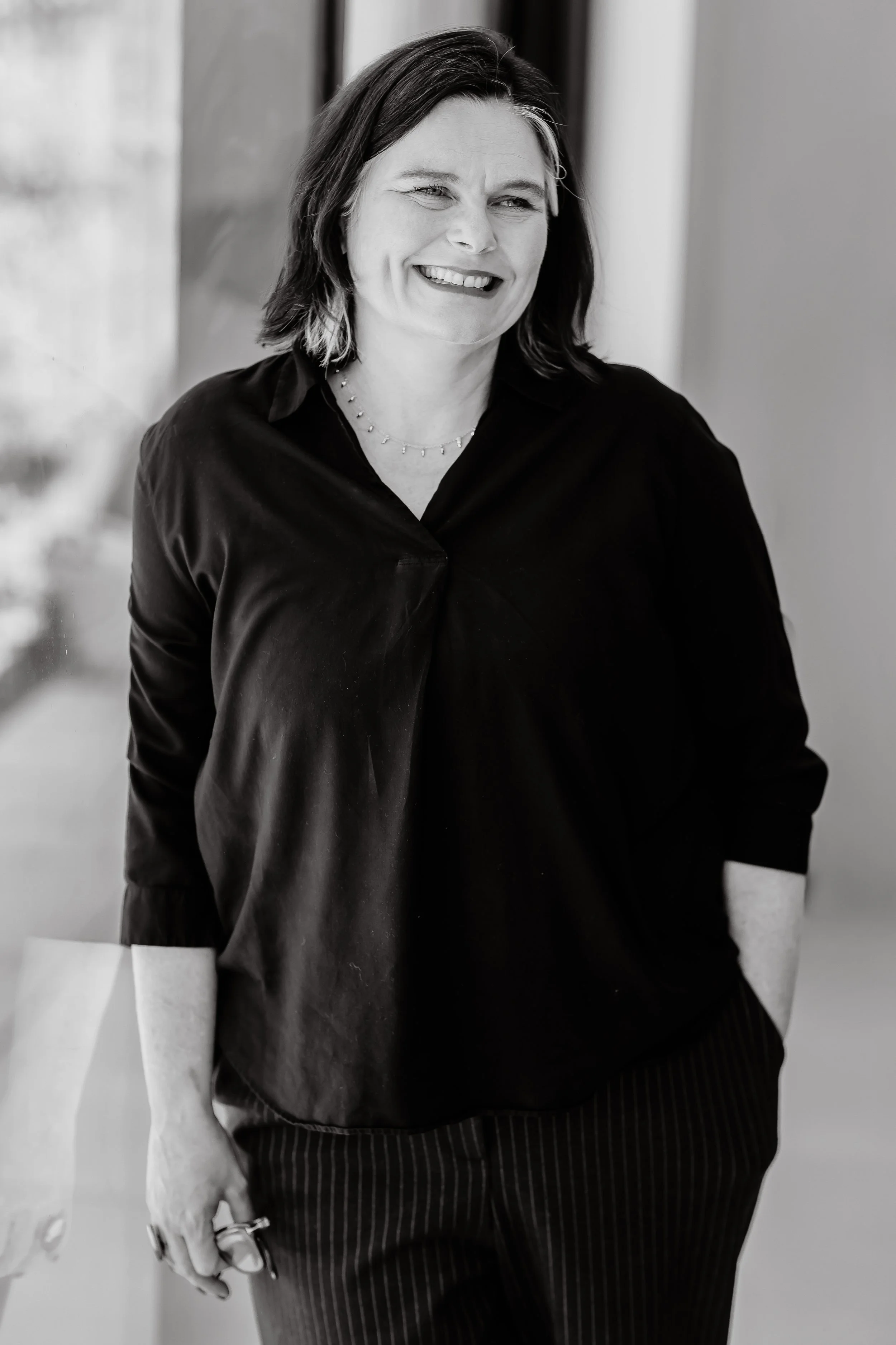 Black-and-white photo of a woman, arts and culture consultant, Jane Macpherson, smiling, wearing a dark top with rolled-up sleeves, and a necklace.