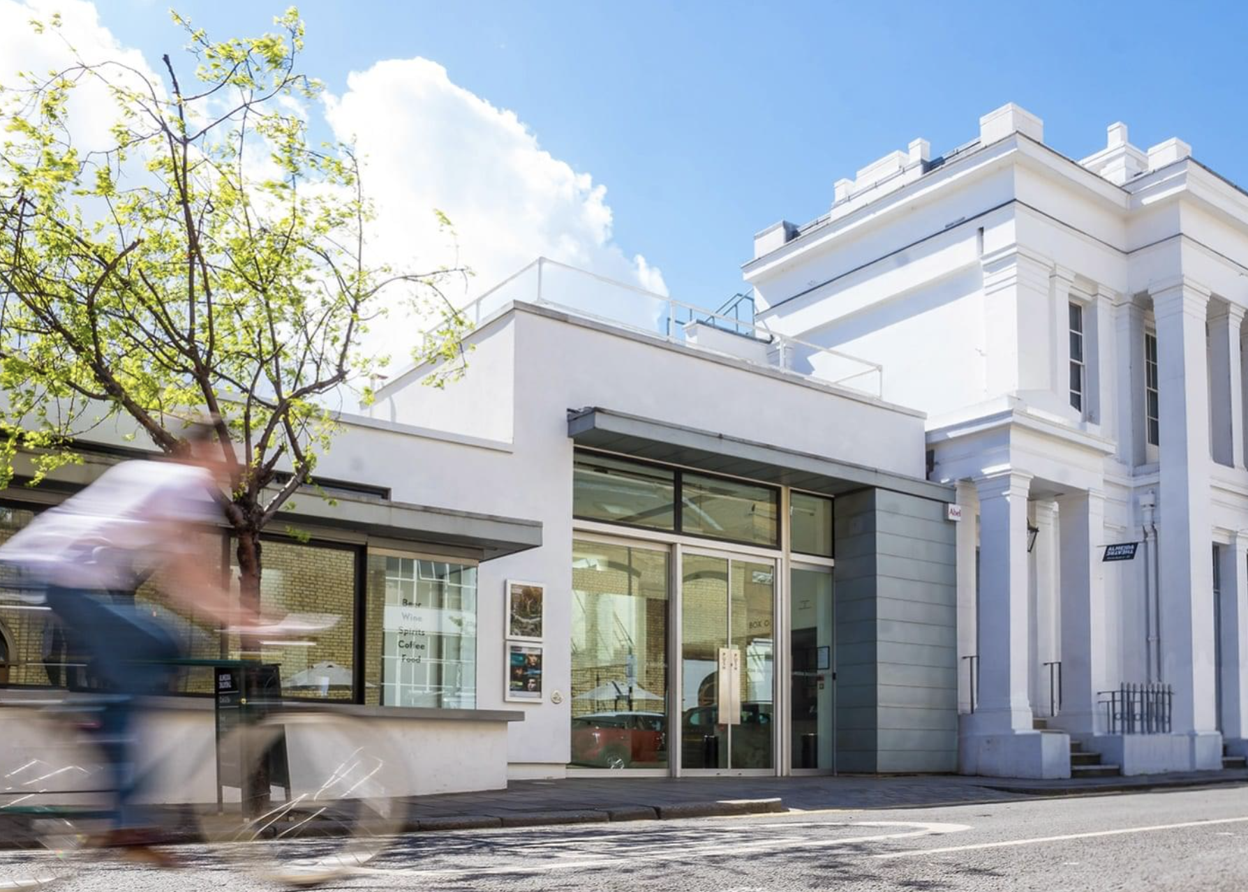 Modern white building with large glass storefront, green and white signage, and a small tree in front. A blurred cyclist passes by on the street under a bright blue sky with fluffy clouds.