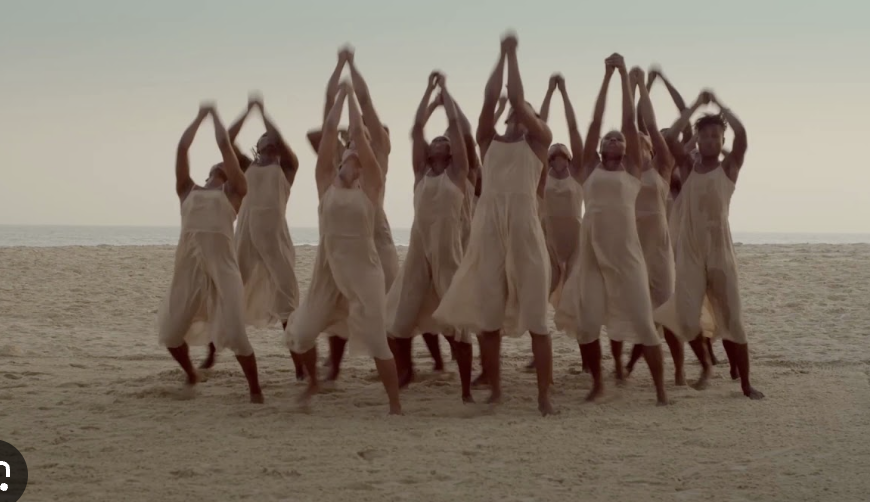 A group of women wearing white dress on a beach, standing with their arms raised above their heads.