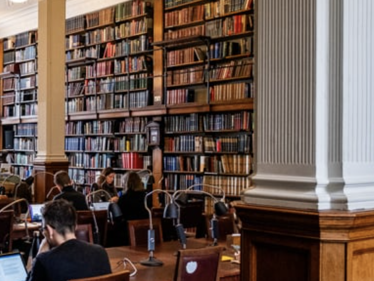 People working or studying at wooden tables in a grand, historic library with tall bookshelves filled with colorful books and large white columns.