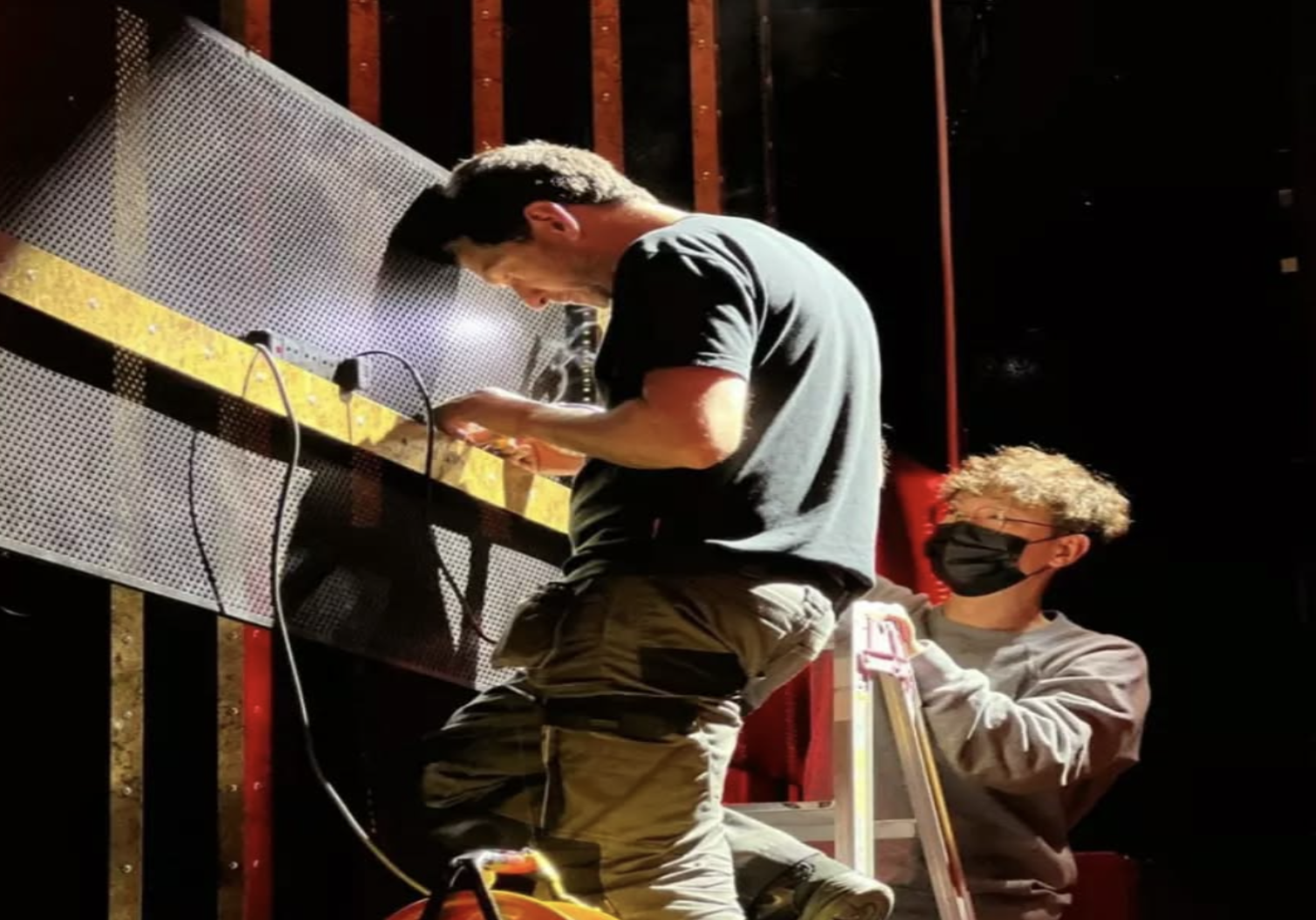 Two young men in a workshop or studio setting. One is working with electronic equipment on a metal workbench, wearing a black T-shirt and camouflage pants. The other, wearing glasses and a black face mask, is holding a ladder and standing behind him,