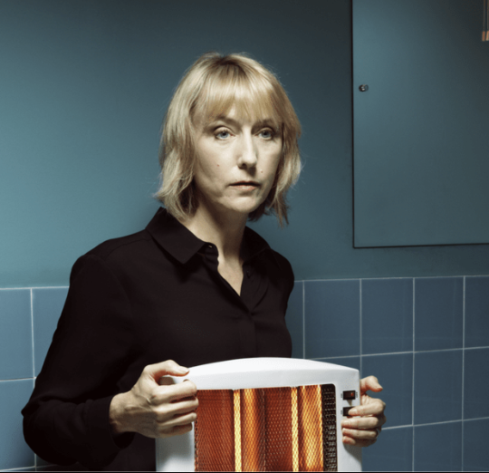 A woman with blonde hair wearing a black shirt holding a space heater in a room with blue walls.