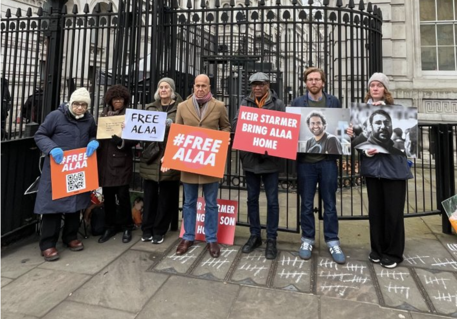 A group of seven people standing behind a black metal fence holding signs and photographs. The signs include messages like '#FREE ALAA', 'FREE ALAA', 'KEIR STARMER BRING ALAA HOME', and there are portraits of a man on some signs. The individuals are 