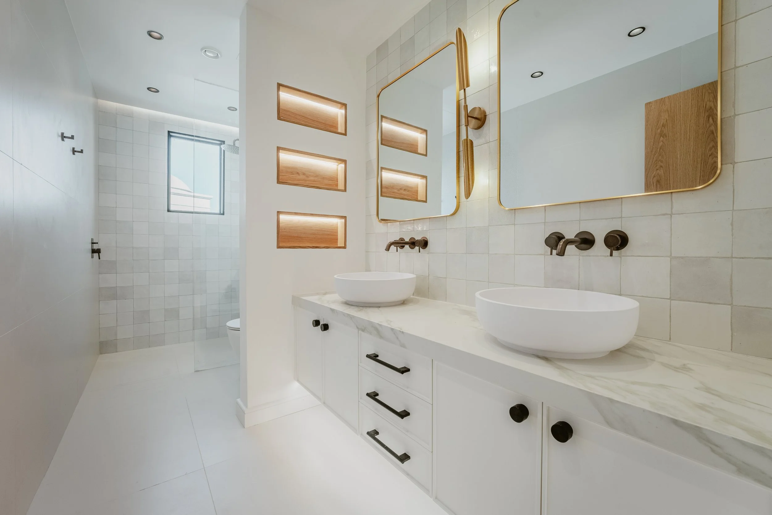 Renovated Al Mahra villa bathroom with double vanity, brass mirrors, wall niches and neutral tile finishes.