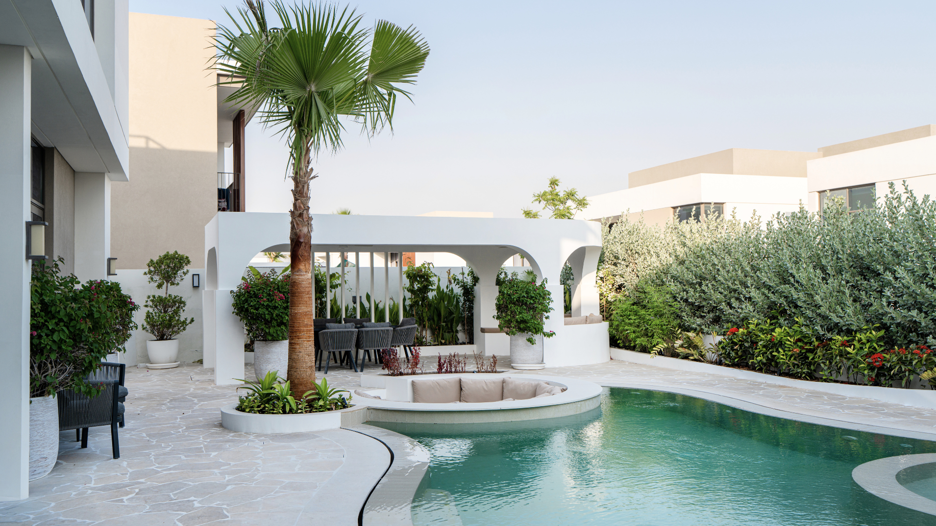 Organic villa pool with integrated seating, palm tree and covered dining pavilion in Tilal Al Furjan.
