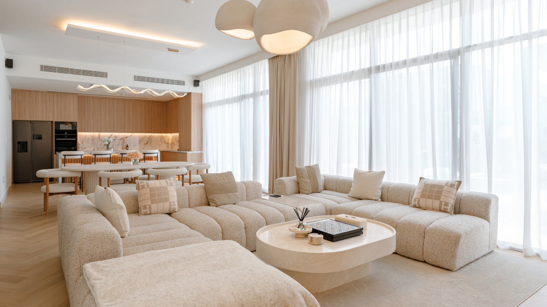 Open-plan villa living room with neutral sectional sofa, bespoke kitchen and floor-to-ceiling windows.