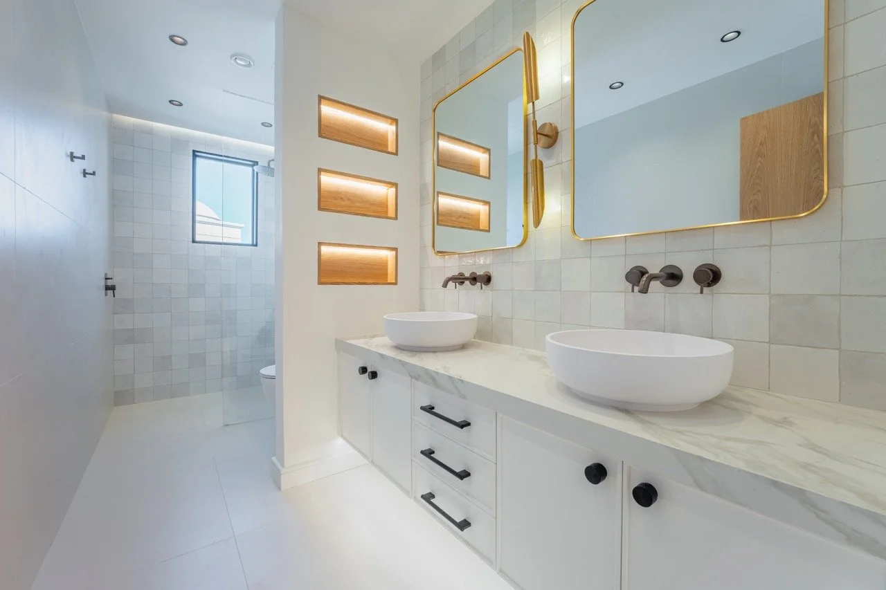 Renovated bathroom with double vanity, brass mirrors, wall niches and neutral tile finishes.