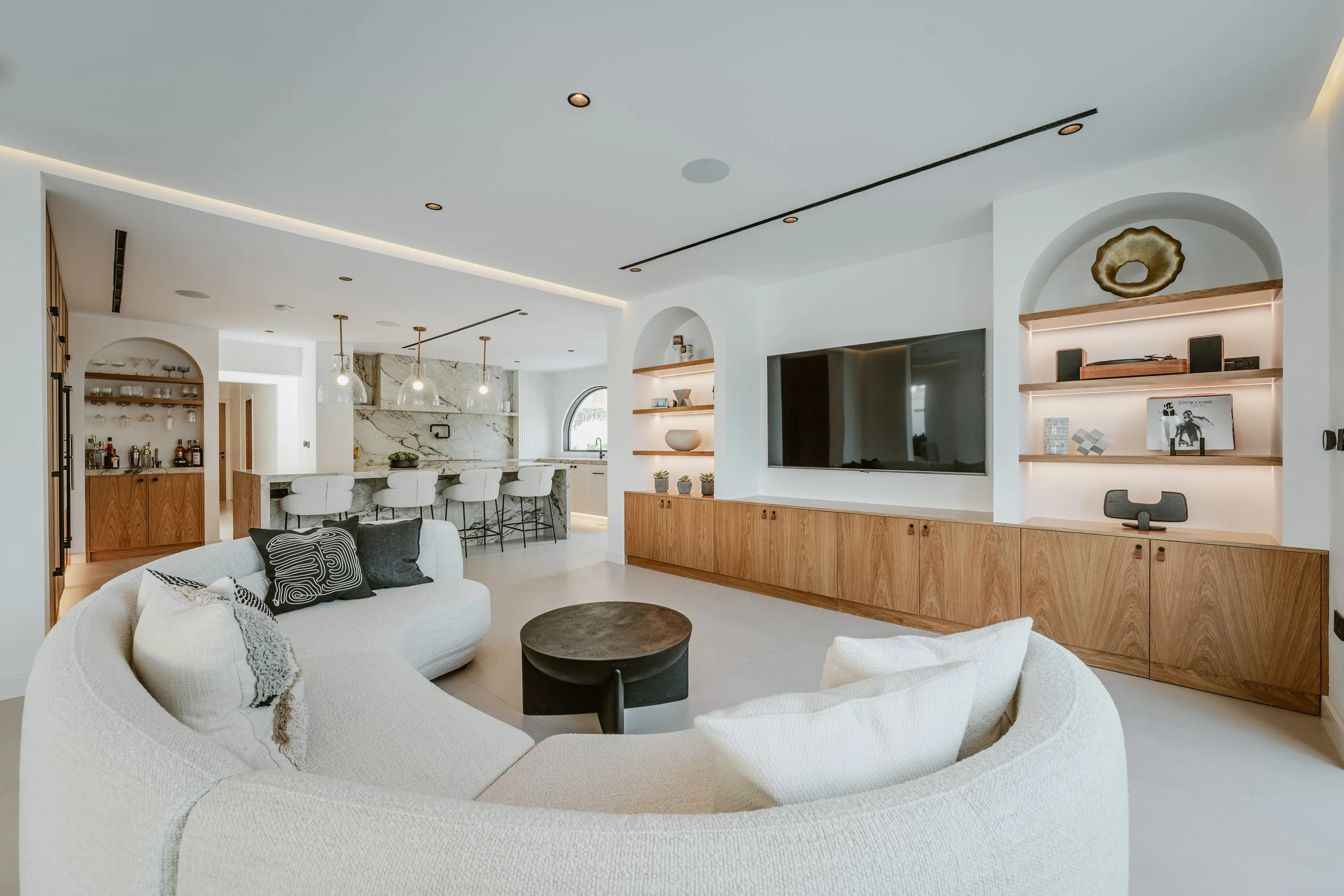 Al Mahra Arabian Ranches villa living room with curved sofa, custom media wall, arched shelving and open-plan kitchen.