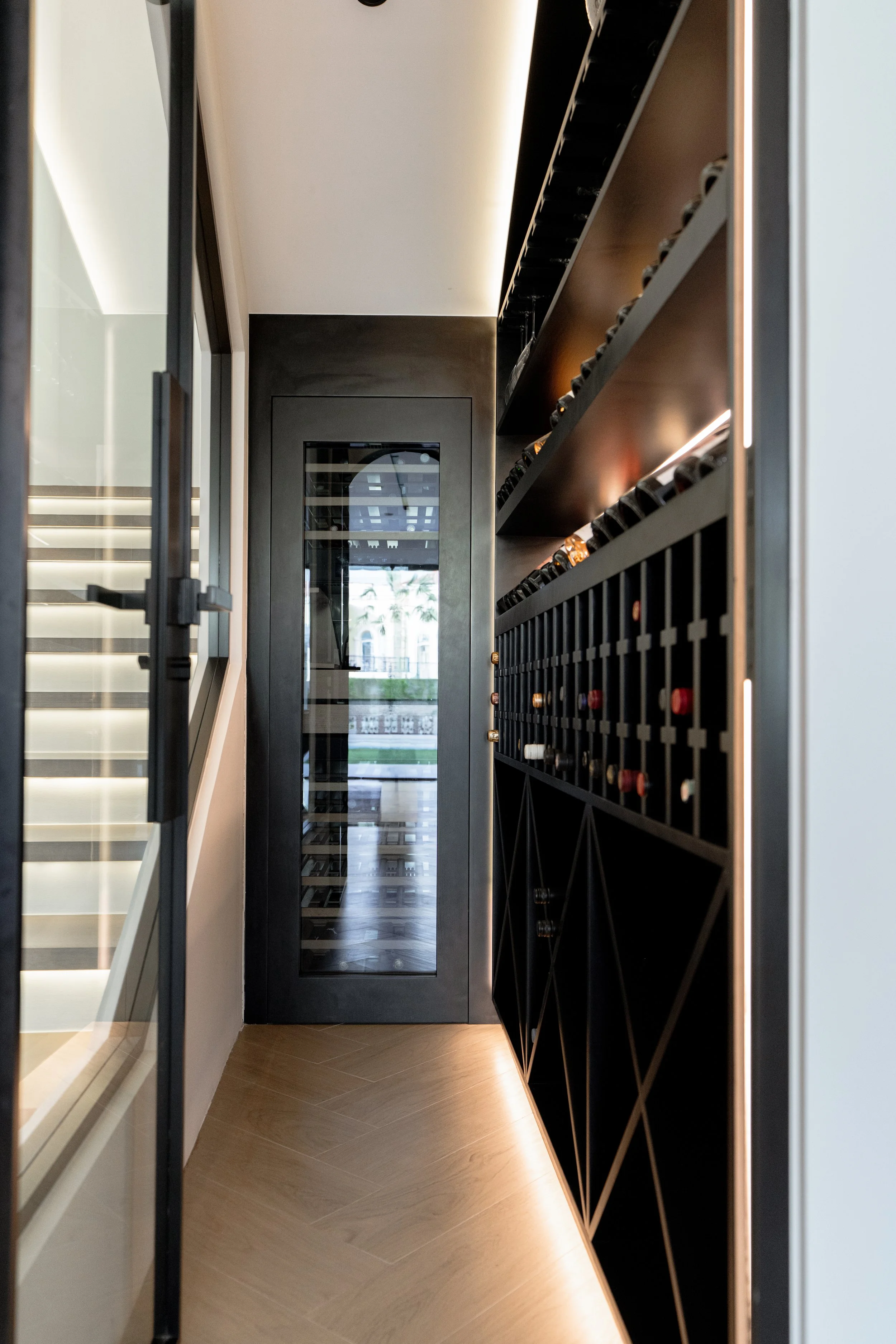 Custom wine storage joinery with integrated lighting and glass door beside a villa staircase