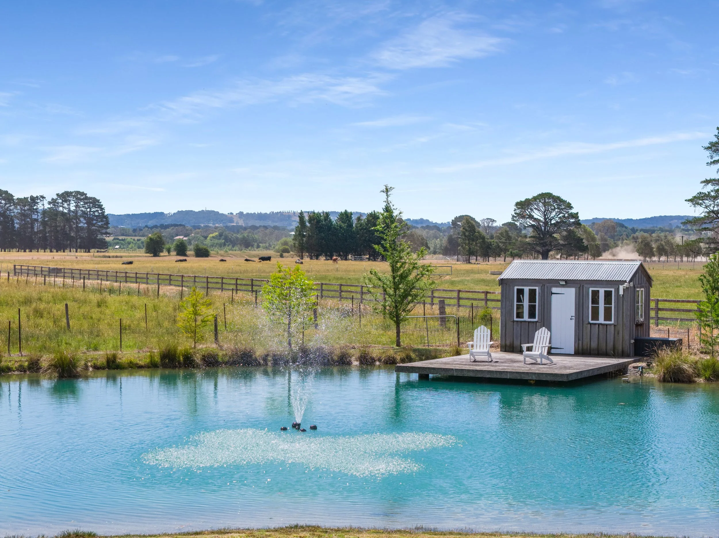 A pond with a fountain, a wooden shed with two Adirondack chairs on a deck, and a rural landscape with grassy fields, trees, and a fence under a blue sky.