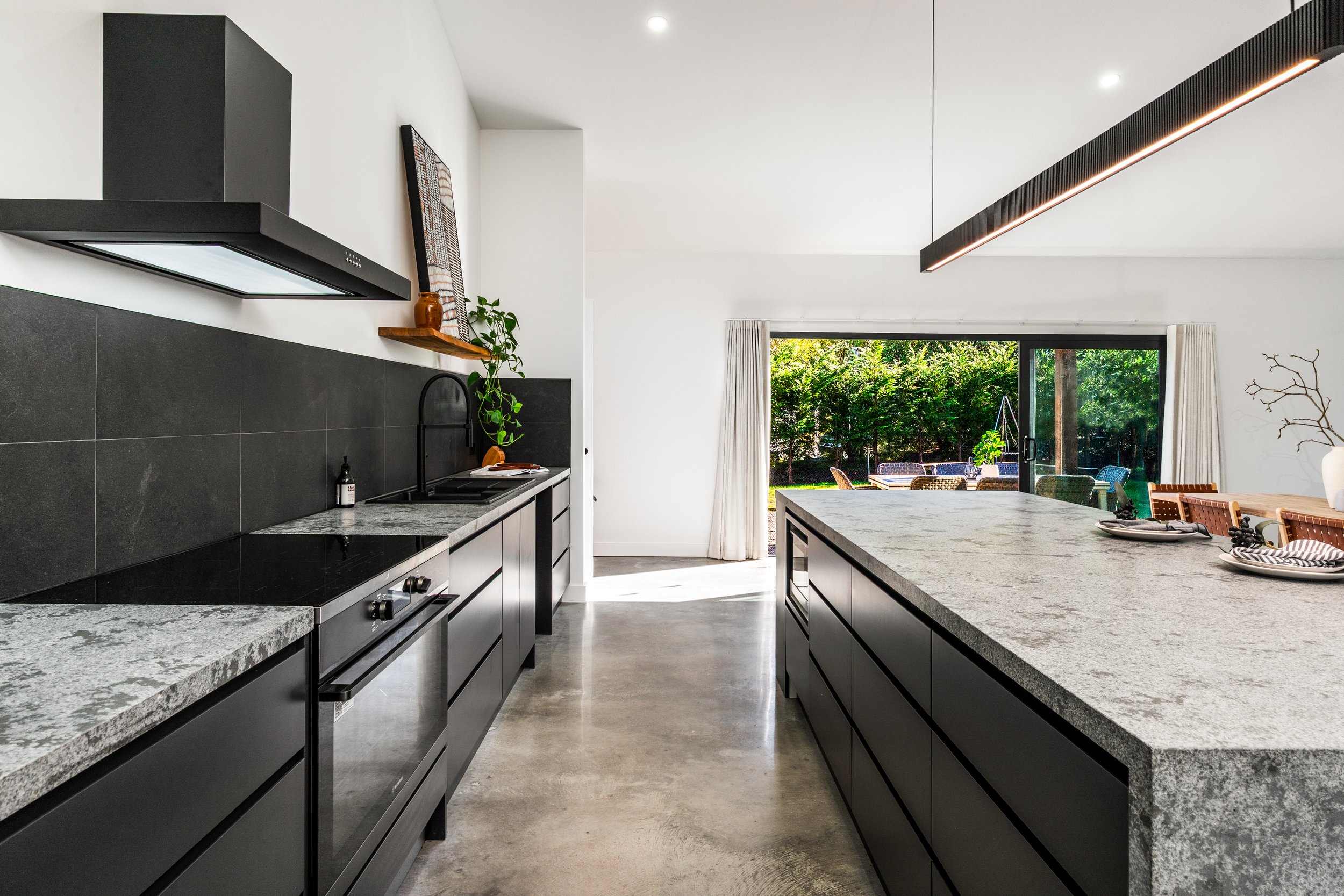 Modern kitchen with black cabinets, gray marble countertops, a large island, and a sliding glass door opening to a garden outdoor seating area.