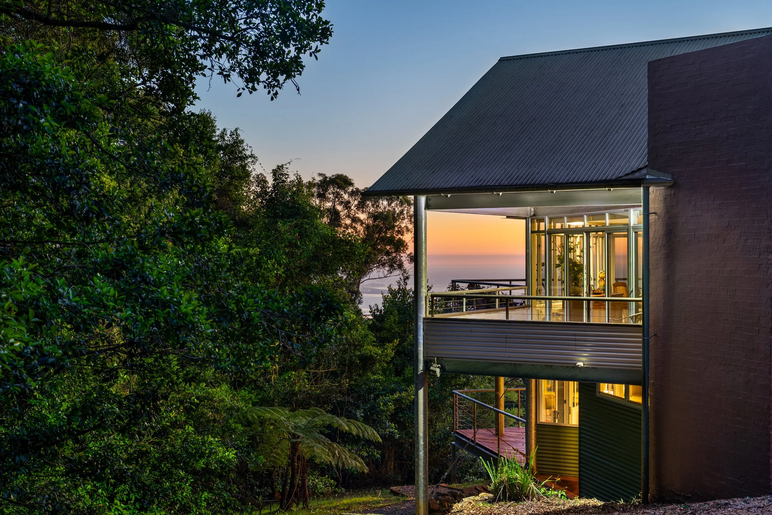 A house with a metal roof and multiple balconies lit from inside, surrounded by lush green trees during sunset.