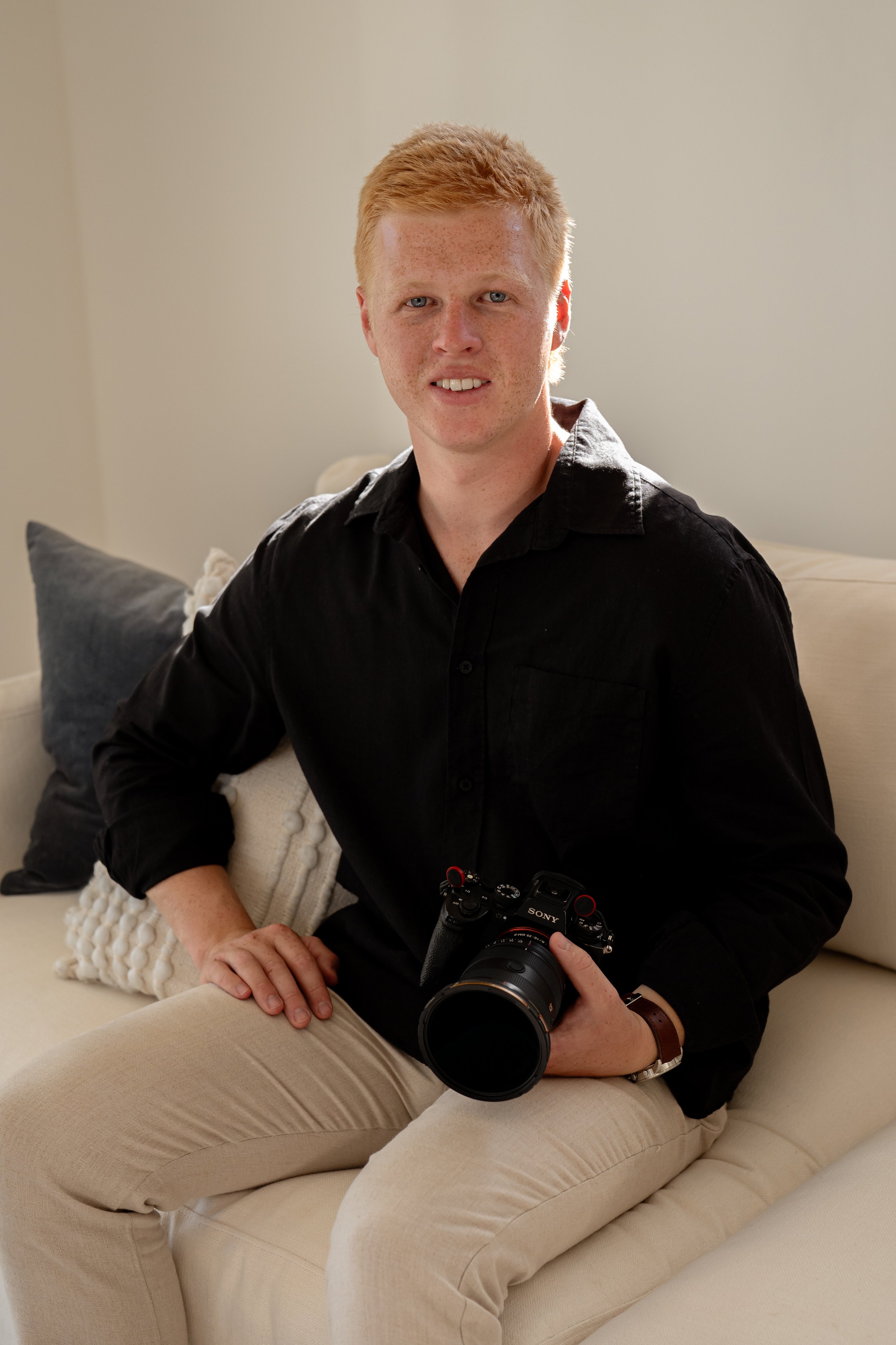 A young man with curly red hair, wearing a black shirt and beige pants, sitting on a cream-colored couch with a camera in his hand, smiling at the camera.