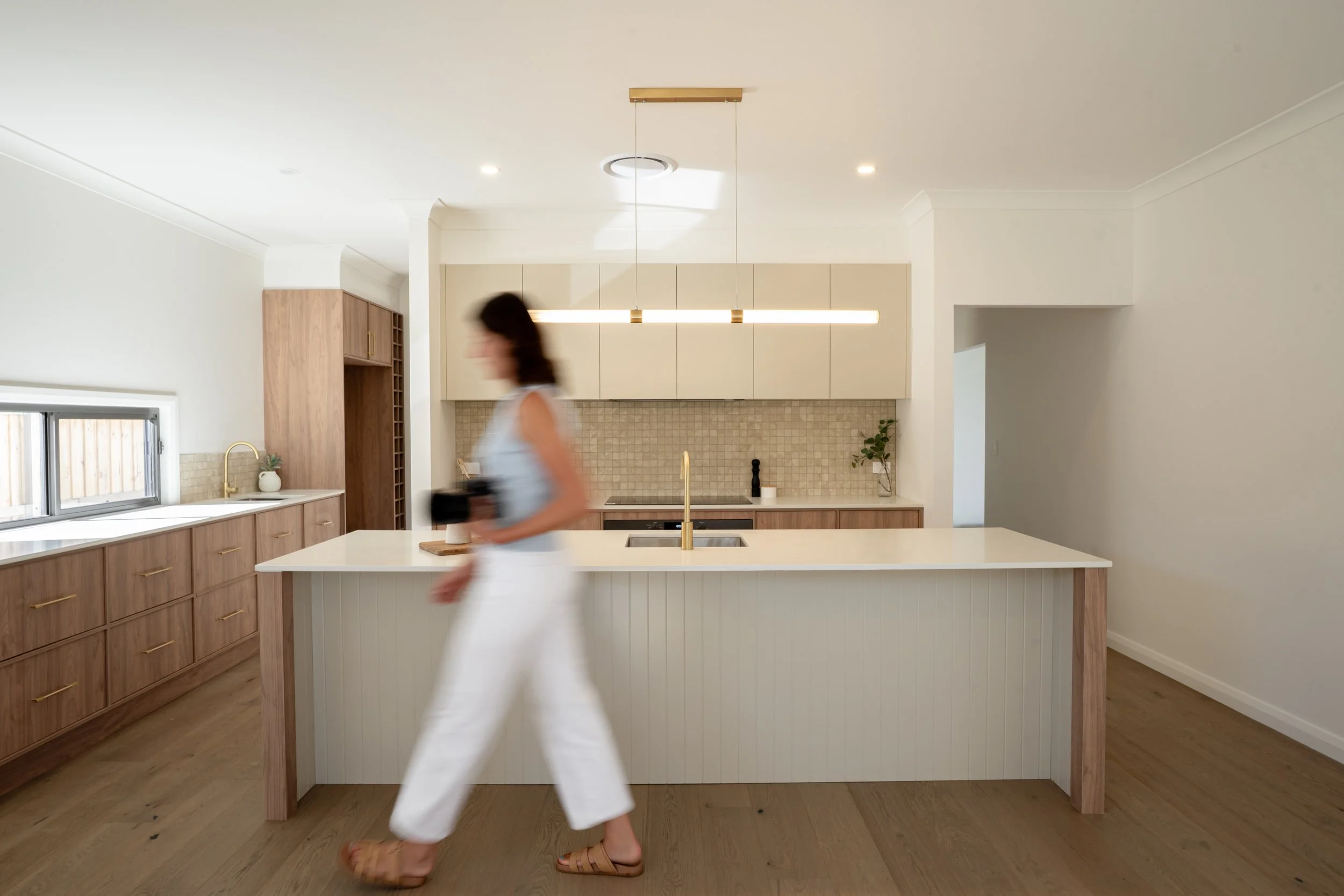 A woman walking in a modern kitchen holding a camera, with wooden cabinets, a kitchen island, and a tiled backsplash.
