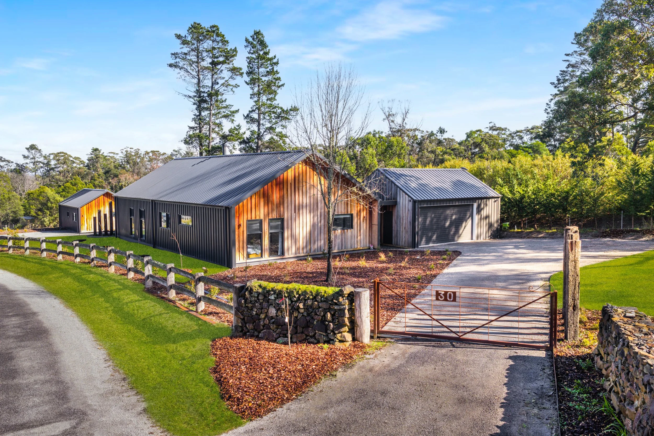 A modern house with a combination of wood and black siding, metal roof, surrounded by green grass and trees, with a gravel driveway and a gate marked with the number 30.