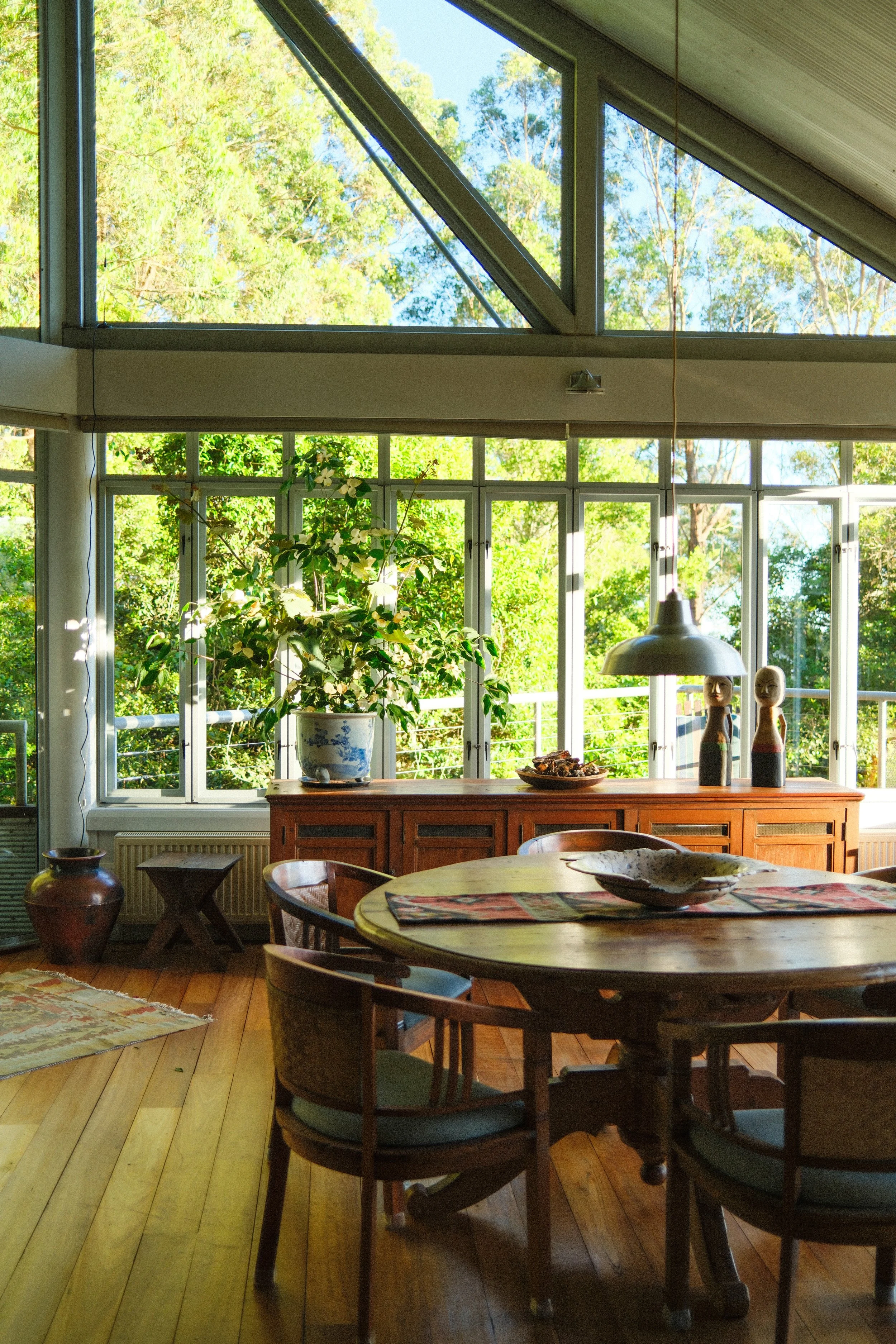 Bright dining room with large windows showing green trees outside, furnished with a round wooden table, chairs, a wooden sideboard, a potted plant, and decorative items.