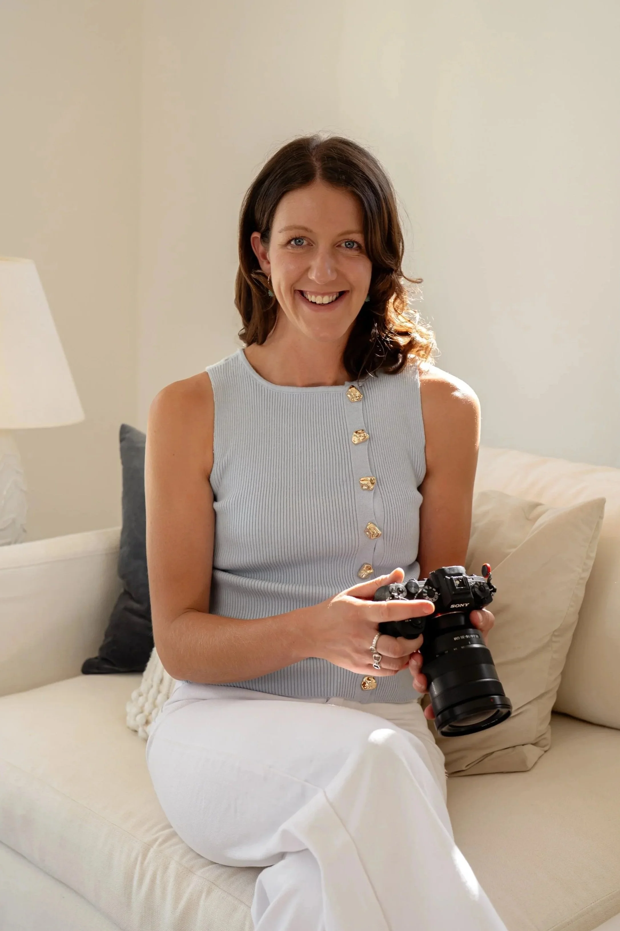 A woman with shoulder-length brown hair, smiling, holding a Sony camera, sitting on a light-colored couch in a bright room with a lamp and pillows.