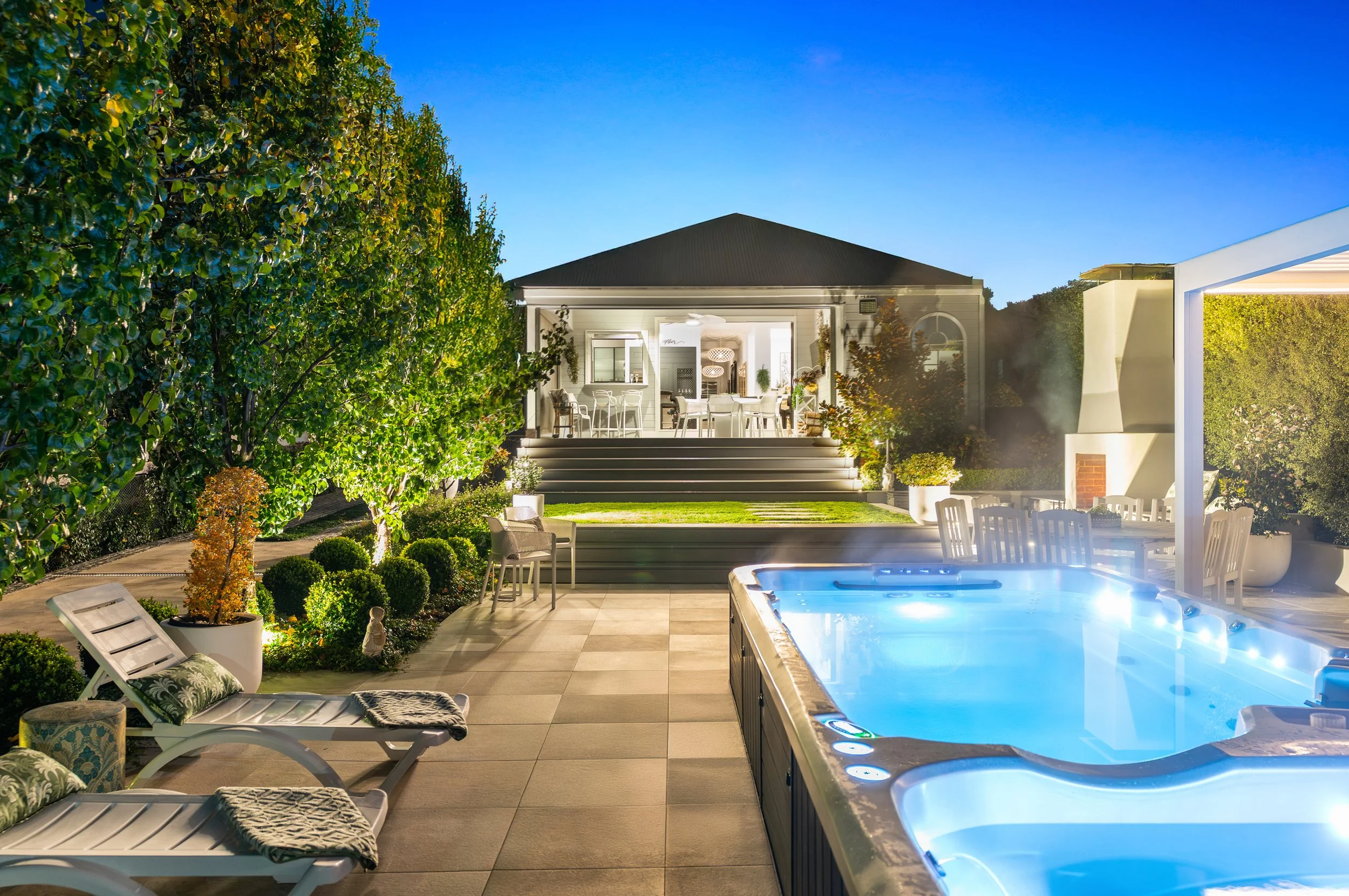 A backyard at dusk with a hot tub, lounge chairs, a dining table, and a house with a lit porch and outdoor kitchen, surrounded by lush green trees and bushes.