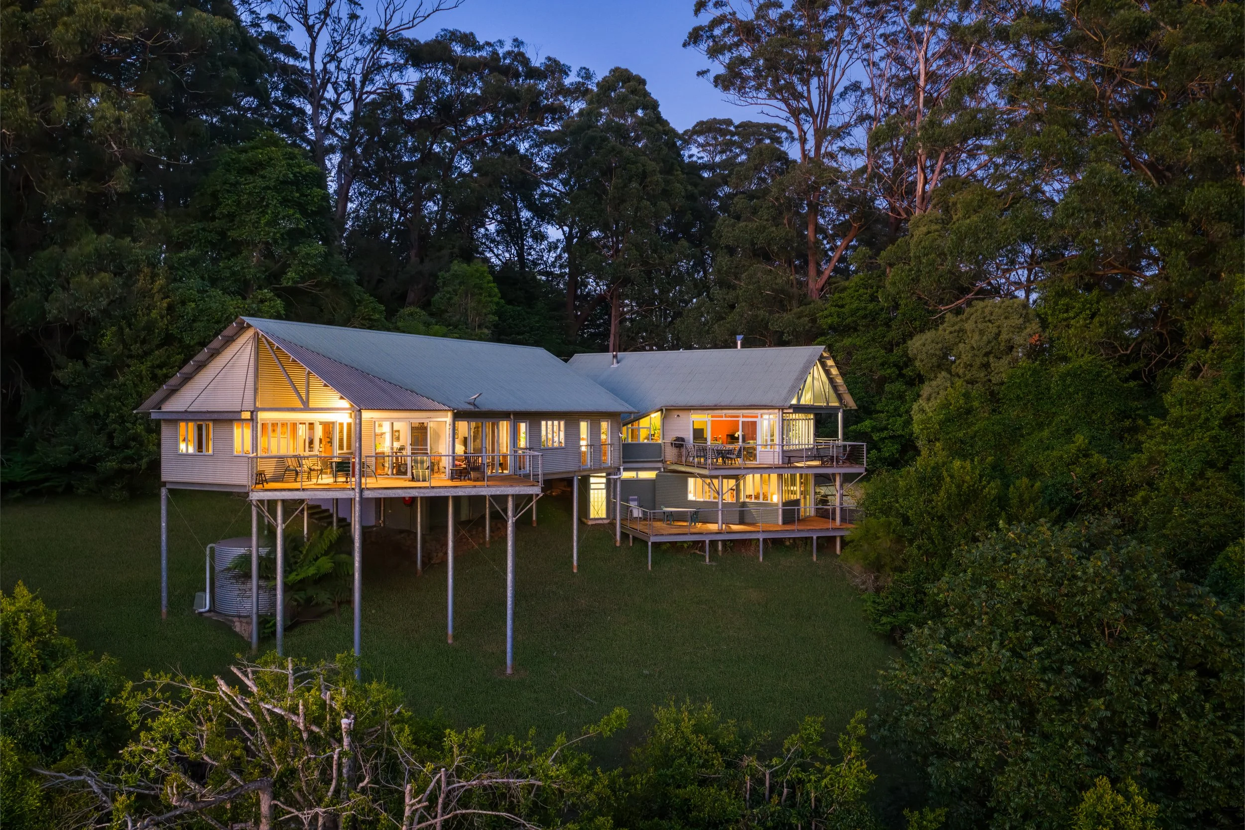 A house built on stilts in a forested area at dusk, with lights on inside, surrounded by tall trees.
