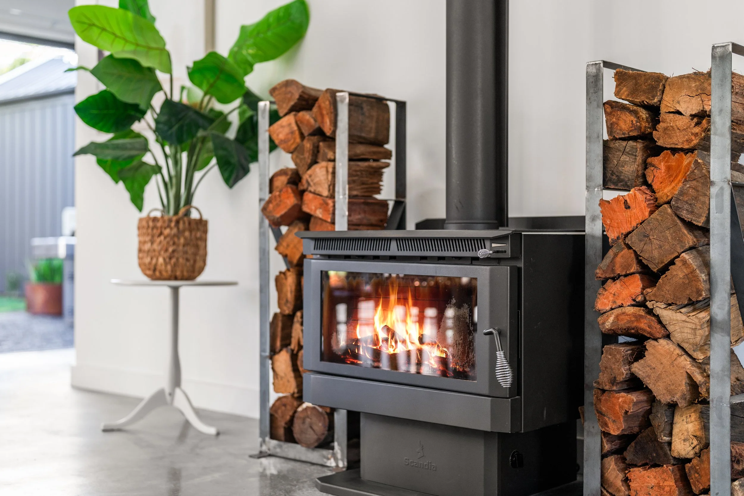 A modern gray wood stove with a fire burning inside, flanked by stacked firewood on metal racks, next to a potted plant on a small white table, in a bright room with a gray floor and wall.